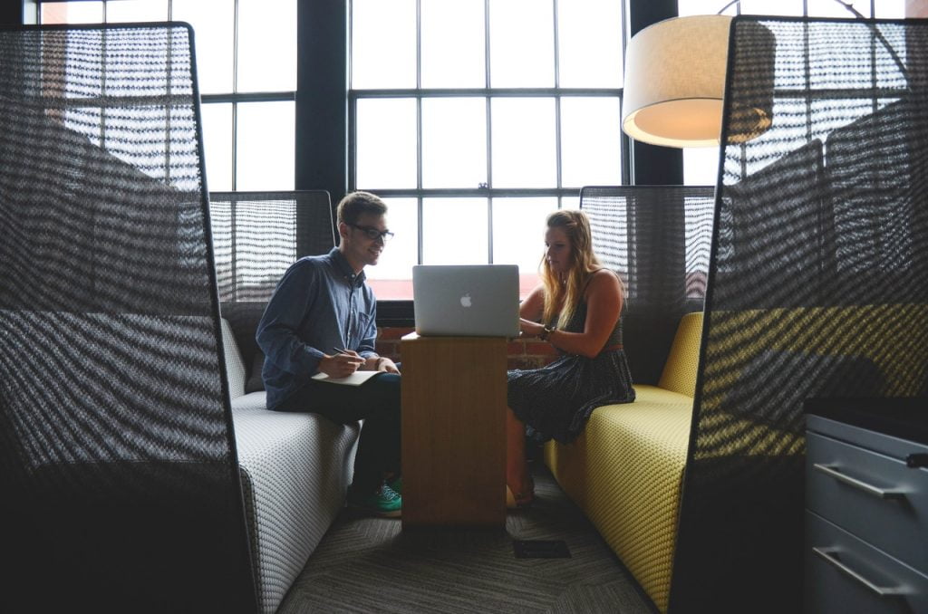 a man and a woman sitting at a desk in a room with a laptop and a window